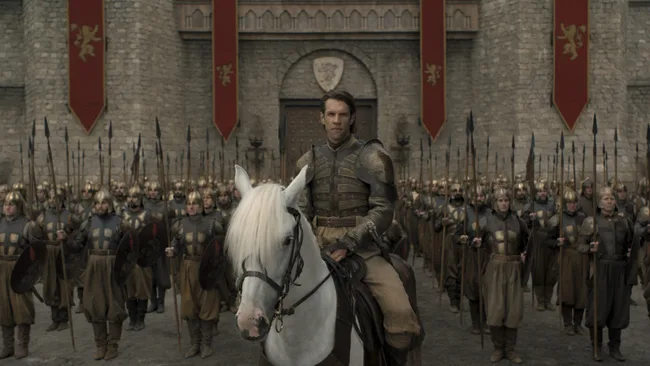 A man in armor rides a white horse in front of a formation of soldiers holding spears, standing outside a castle gate.