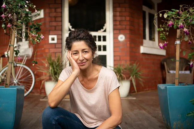 Woman sitting on a porch with her hand on her cheek, surrounded by plants and a bicycle in the background.