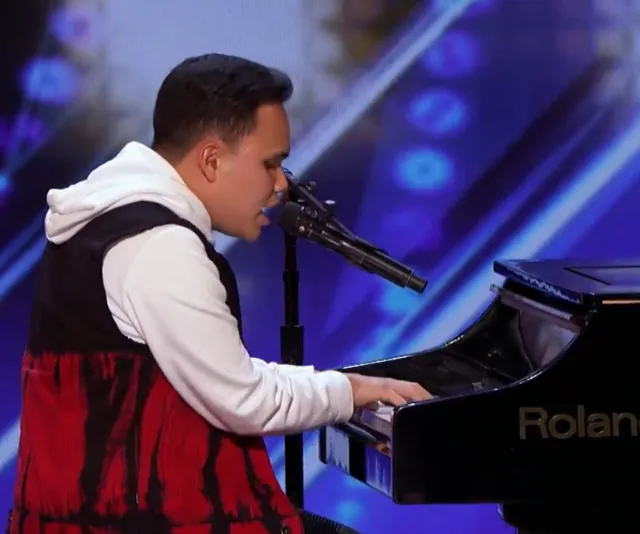 Musician playing piano on stage, wearing a hoodie, with a microphone nearby; vibrant blue background.