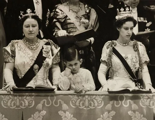 Young boy looking bored at a formal event, flanked by two elegantly dressed women in regal attire.