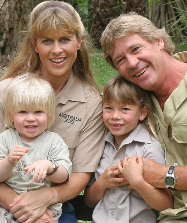 A smiling family of four sitting outdoors, wearing khaki shirts, with trees in the background.