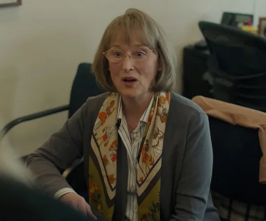 A woman with glasses and a floral scarf sits inside an office setting.