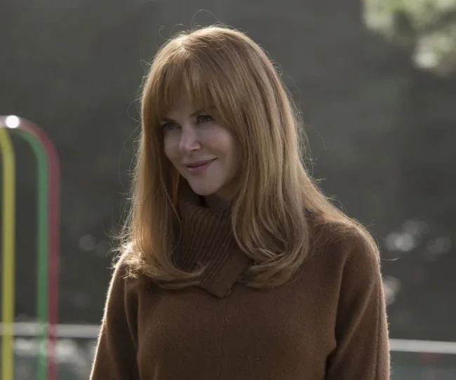 A woman with long red hair and a brown turtleneck smiles outdoors.