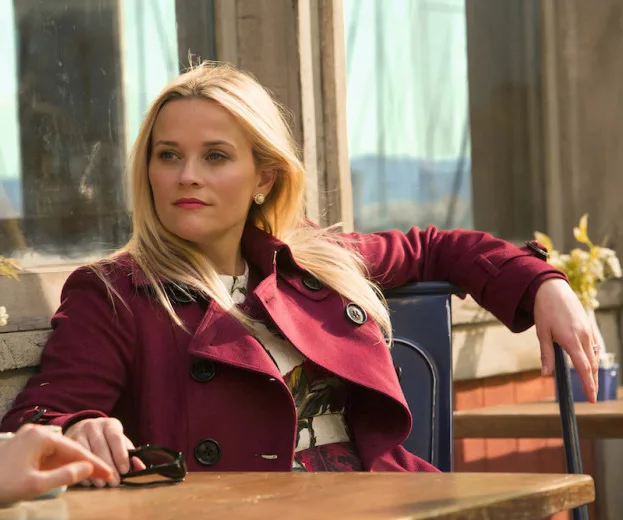 A woman in a burgundy coat sits at an outdoor cafe table, looking away thoughtfully.