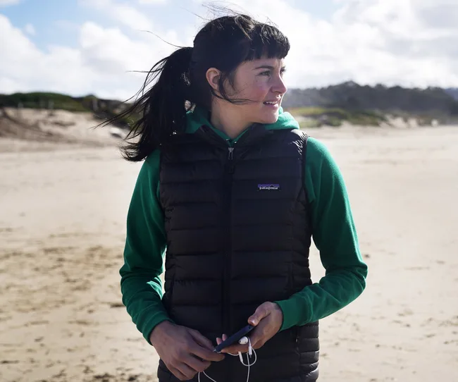 Young woman in a green hoodie and black vest smiles, holding a phone, standing on a sunny, windy beach.