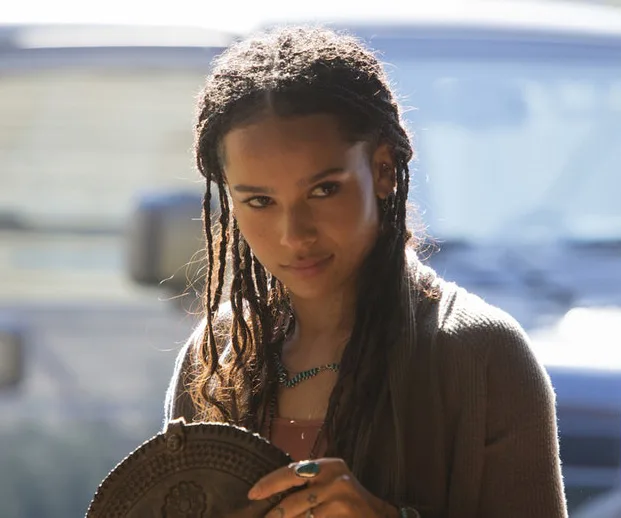 A person with braided hair holding a circular object, standing outside with a car in the background.