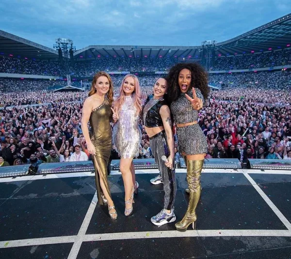 Spice Girls pose on stage before a large cheering crowd at an outdoor stadium concert.