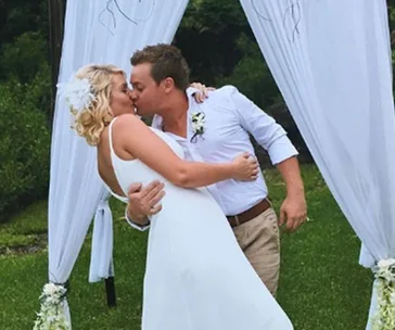 Couple kissing under an outdoor wedding arch, surrounded by greenery.
