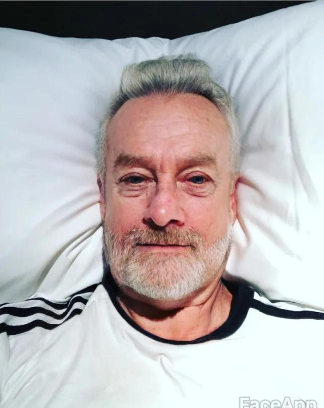 Person with gray beard lying on a pillow, wearing a black and white shirt, smiling slightly.