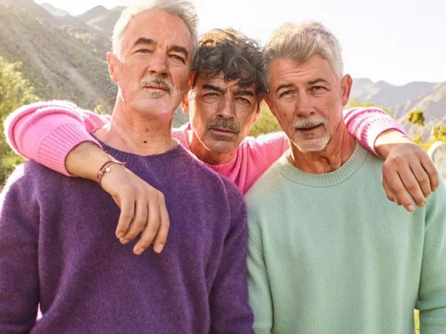 Three older men in colorful sweaters pose together outdoors in a scenic landscape with mountains.