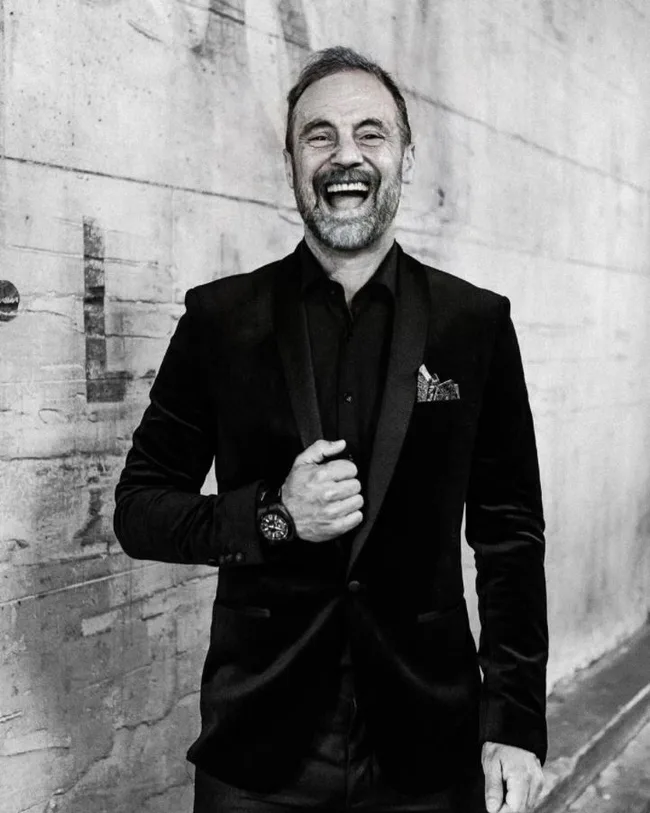 Man in a black suit laughing, standing against a textured wall, wearing a wristwatch.