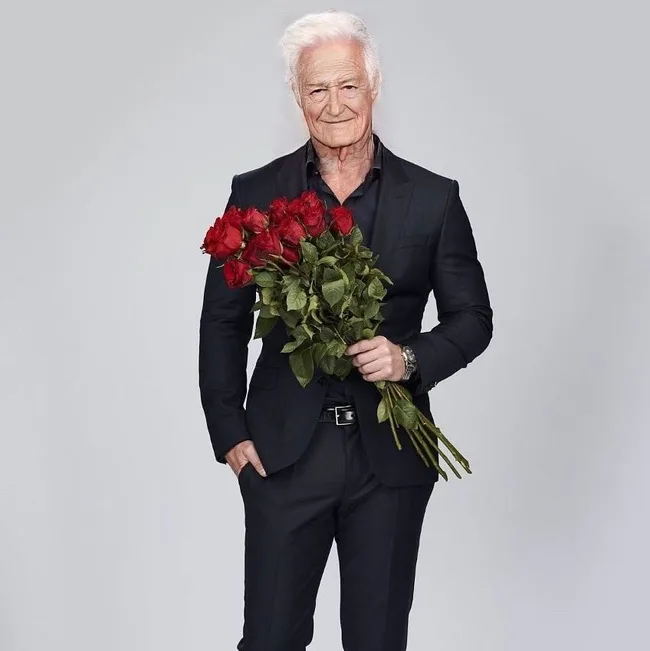 Old man in a black suit holding a bouquet of red roses, smiling slightly against a gray background.