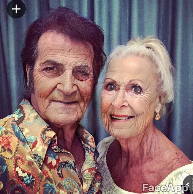 Elderly couple smiling for selfie, edited using FaceApp, with blue curtain background.