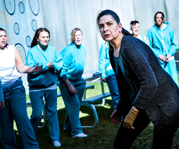 A tense scene from Wentworth Season 7, featuring women in prison uniforms appearing shocked behind a determined character.
