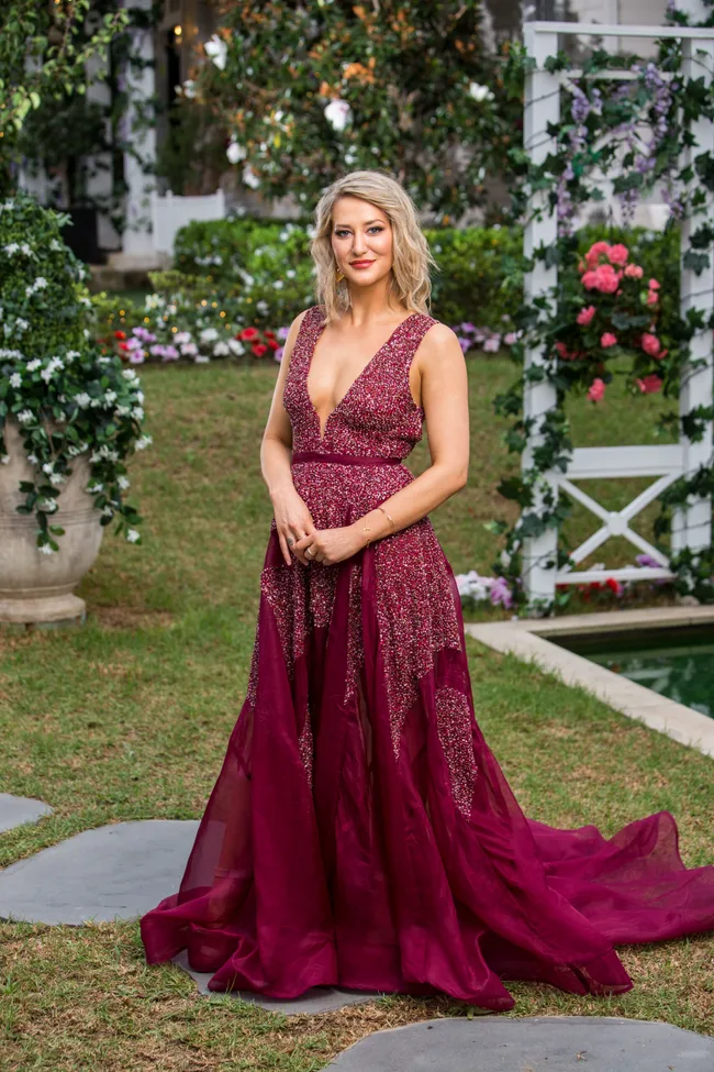 A woman in a red gown stands in a garden, with flowers and greenery as a backdrop.