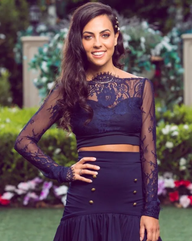 Woman in a navy lace dress, standing in a garden with flowers in the background, smiling at the camera.
