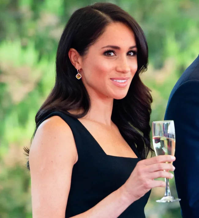 Woman in a sleeveless black dress holding a glass of champagne, smiling with a blurred green background.