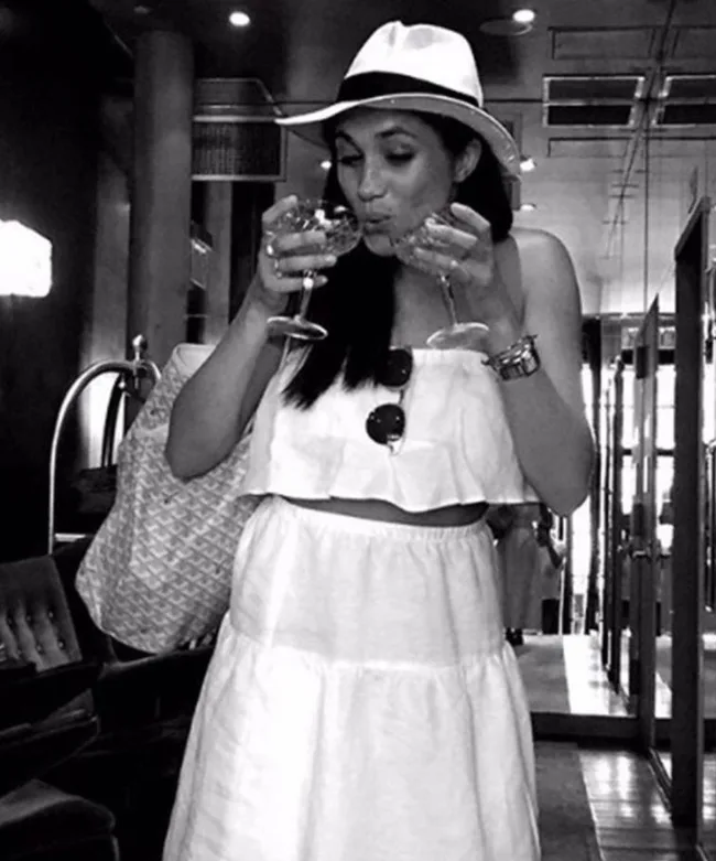 Woman in a white outfit and hat holds two glasses, playfully drinking in a trendy indoor setting.