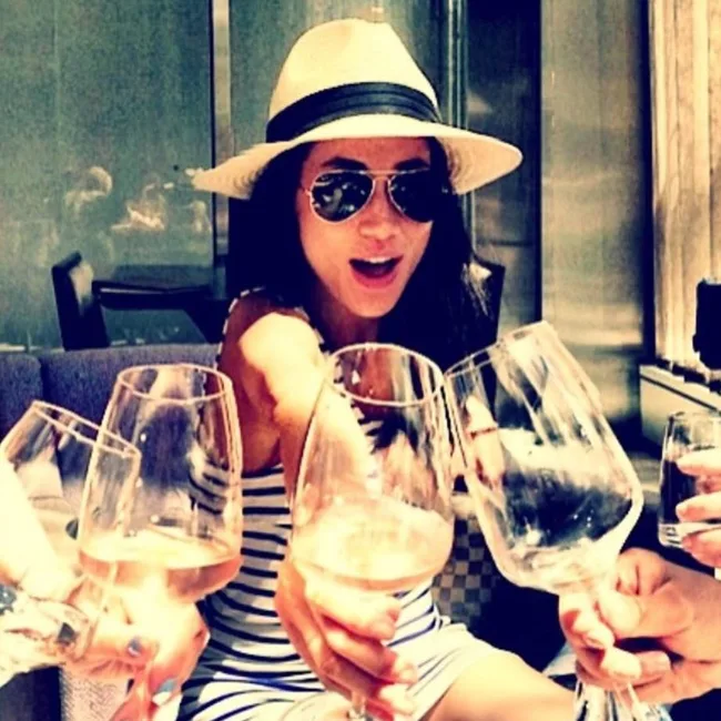 Young woman in a striped dress and hat clinks wine glasses with friends, smiling outdoors.