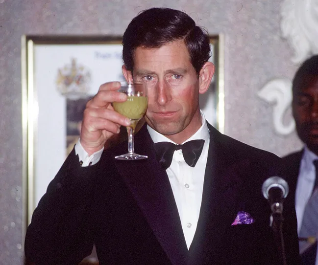 A man in a tuxedo raises a glass in a toast, with a microphone in front of him and another person blurred in the background.