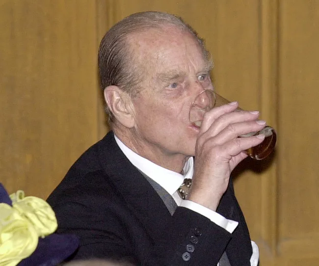 An older man in a suit drinks from a glass at an event.