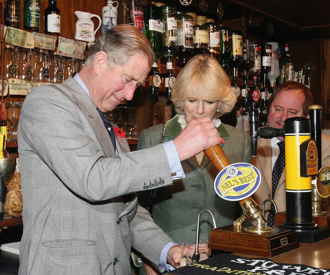 A man and woman pour a drink at a bar; both are wearing suits. Bottles and decor are in the background.