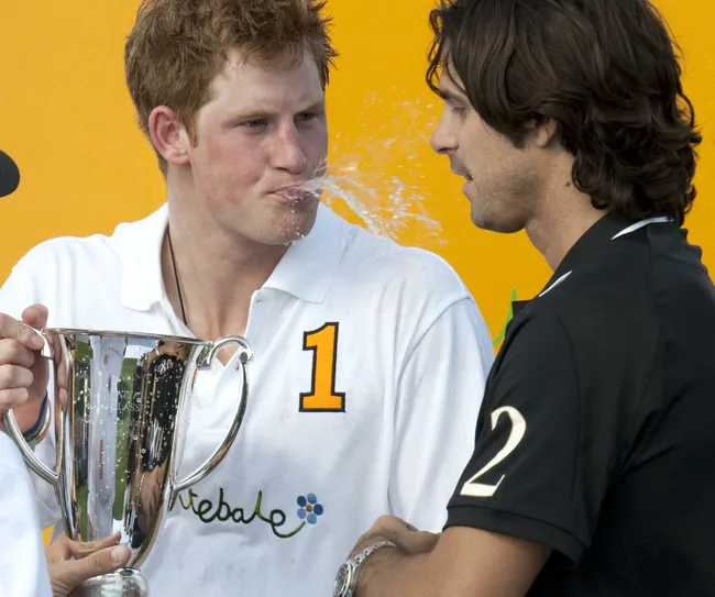 Man in a white shirt with number 1 playfully splashes water from his mouth at another man holding a trophy.