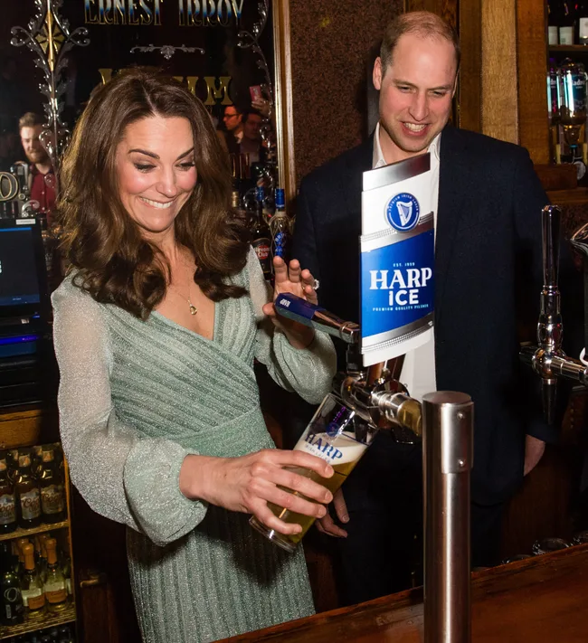 Woman in green dress pouring a Harp lager from a tap, man in suit watches beside her in a pub setting.