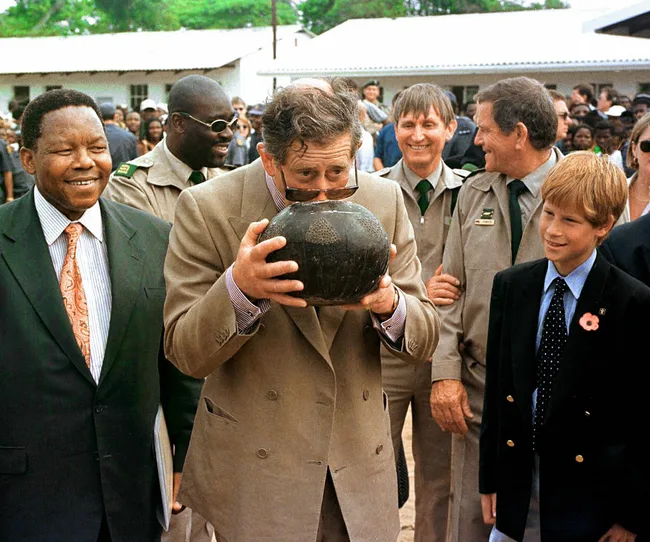 A man drinks from a traditional vessel while a group of people, including a smiling person in a suit, observes outdoors.