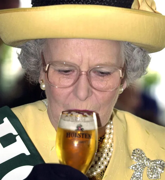 Elderly woman in a yellow outfit with glasses holding a Holsten beer in a glass, wearing a pearl necklace.
