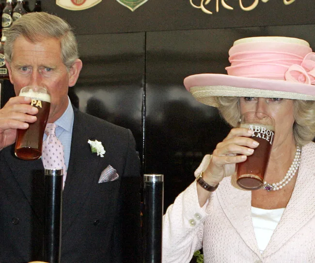 A man in a suit and a woman in a pink hat drink pints of beer in a pub setting.