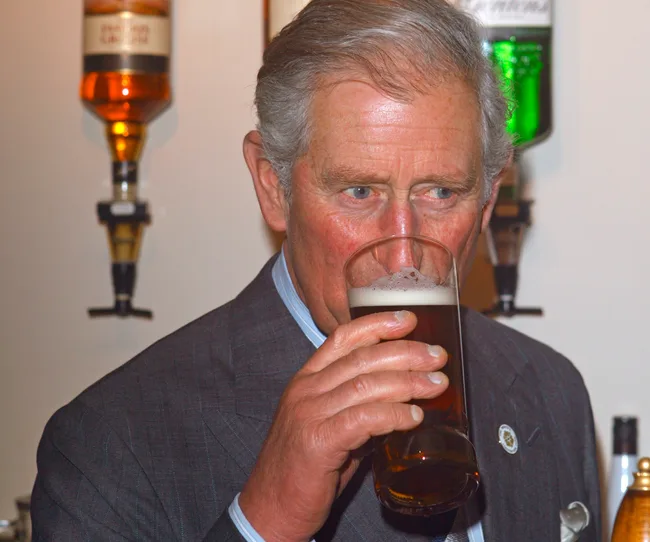Man in a suit drinking a pint of beer, with bottles on wall behind him.