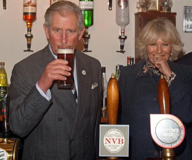 Man in suit drinking beer at a bar with woman smiling beside him; bar backdrop has various bottled drinks displayed.
