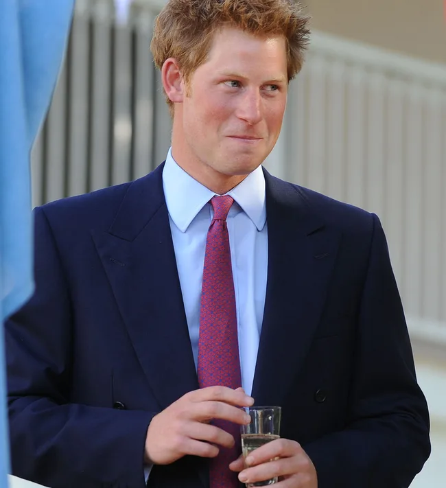 Man in a suit with a red tie, holding a glass drink, standing outdoors.