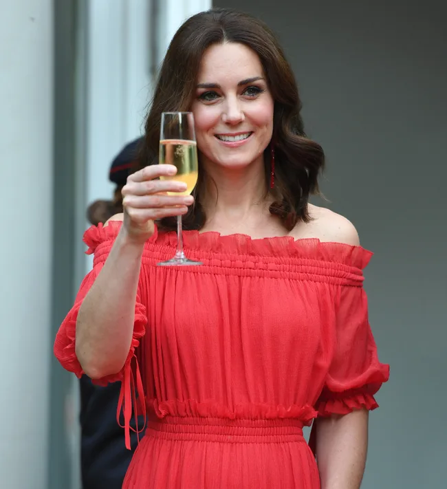 A woman in a red off-shoulder dress raises a glass of champagne, smiling warmly.