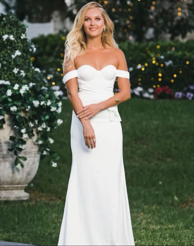 A woman in an elegant white off-shoulder gown stands in a garden, with twinkling lights in the background.