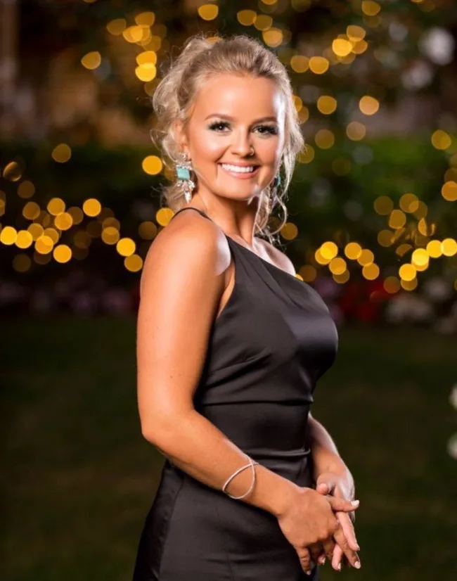 A woman in a black dress poses outdoors at night. Blurry warm lights create a bokeh effect in the background.