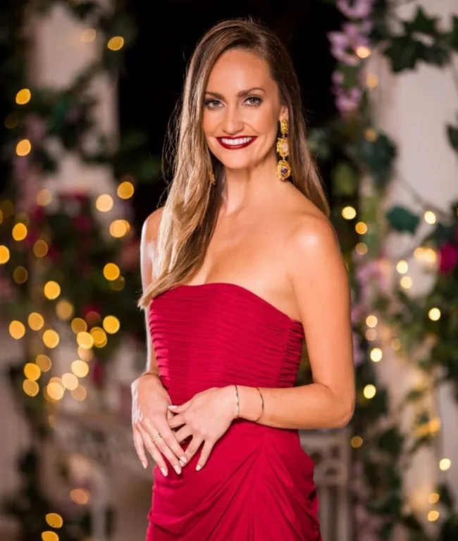 A smiling woman in a red strapless dress with long earrings, standing in front of blurred decorative lights and flowers.