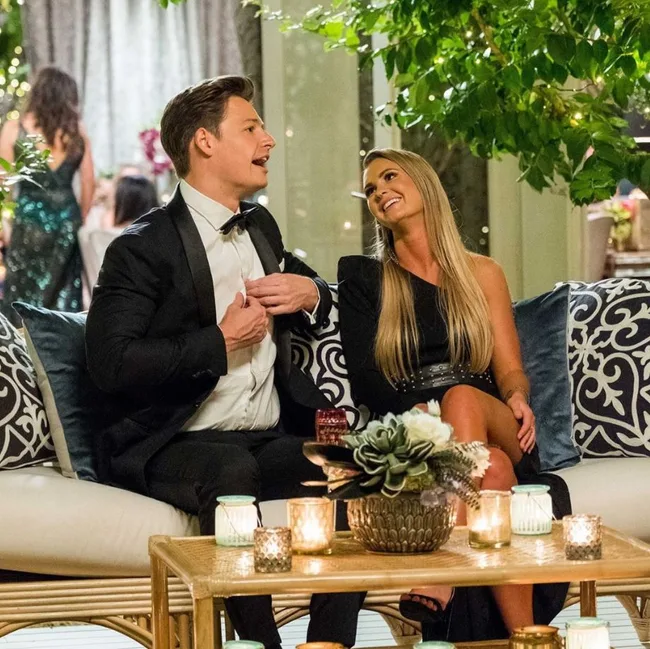 Two people in formal wear sitting on a sofa, surrounded by candles and plants, engaged in conversation at an event.