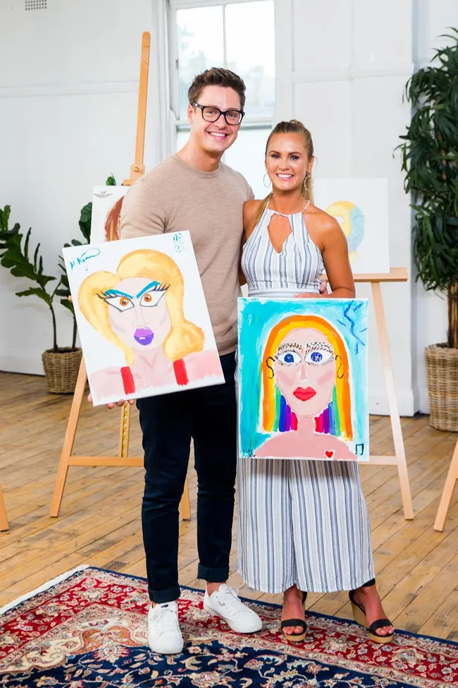 Two people smiling, holding colorful abstract portraits in an art studio setting.