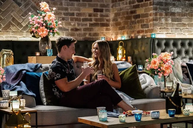 A couple sitting on a cozy sofa, surrounded by flowers and candles, enjoying drinks in a warmly lit setting.