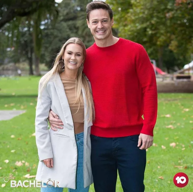 A man in a red sweater with his arm around a woman in a white coat, smiling in a park setting with "The Bachelor" logo.