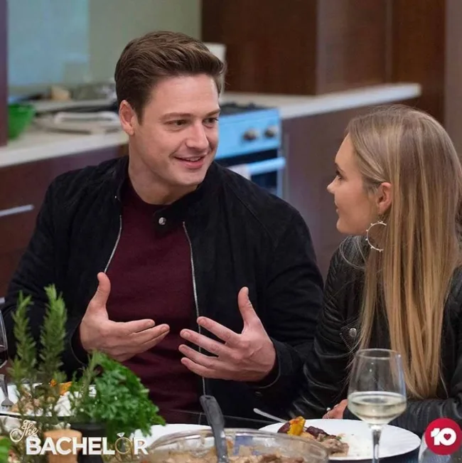 Man and woman talking at a dinner table with "The Bachelor" logo in the corner.