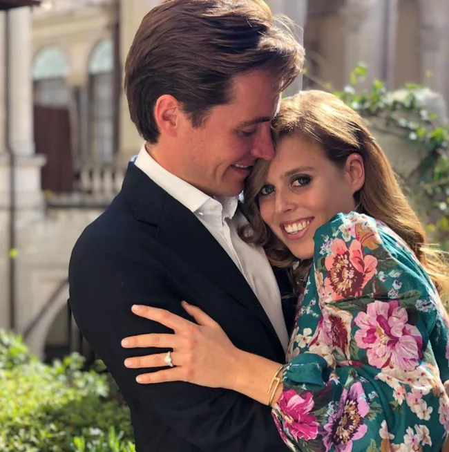 A couple embraces outdoors; the woman wears a floral dress and shows an engagement ring with a smile.