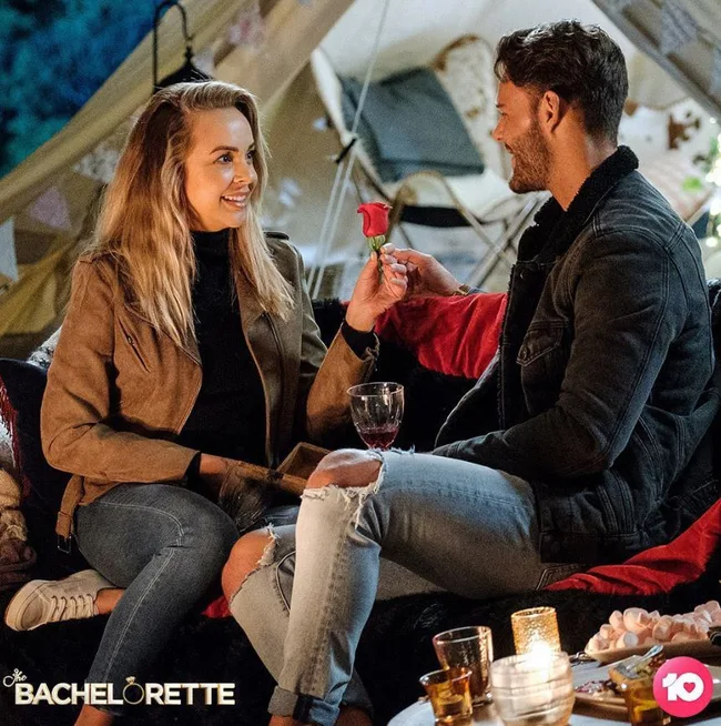 A couple sits in a cozy outdoor setting, with one holding a rose; "The Bachelorette" logo is visible at the bottom.