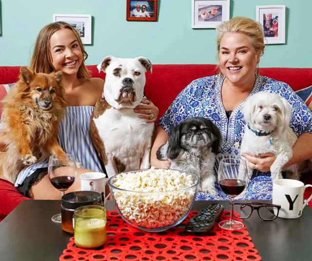 Two women and four dogs sitting on a red couch with wine and popcorn on a table.