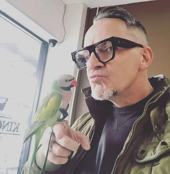 A person wearing glasses makes a face while holding a green parrot on their finger indoors.