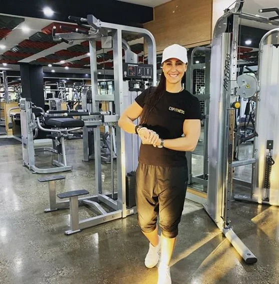 A woman in a black workout outfit and white cap smiling in a gym, surrounded by exercise equipment.