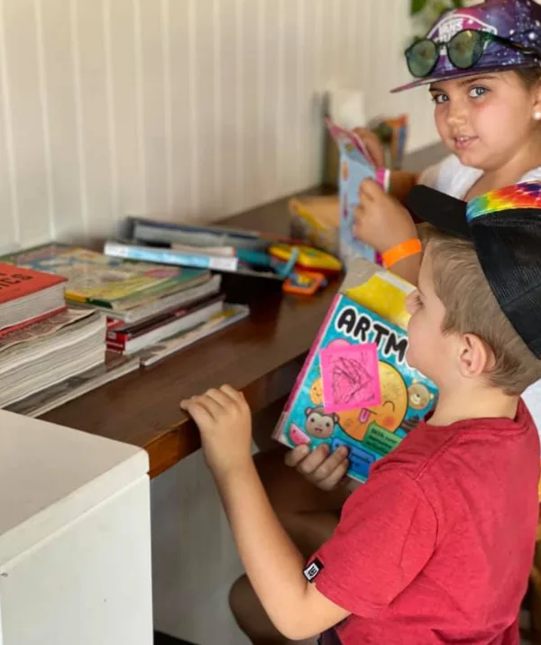 Children looking at children's books and magazines on a wooden shelf, with one holding a colorful activity book.