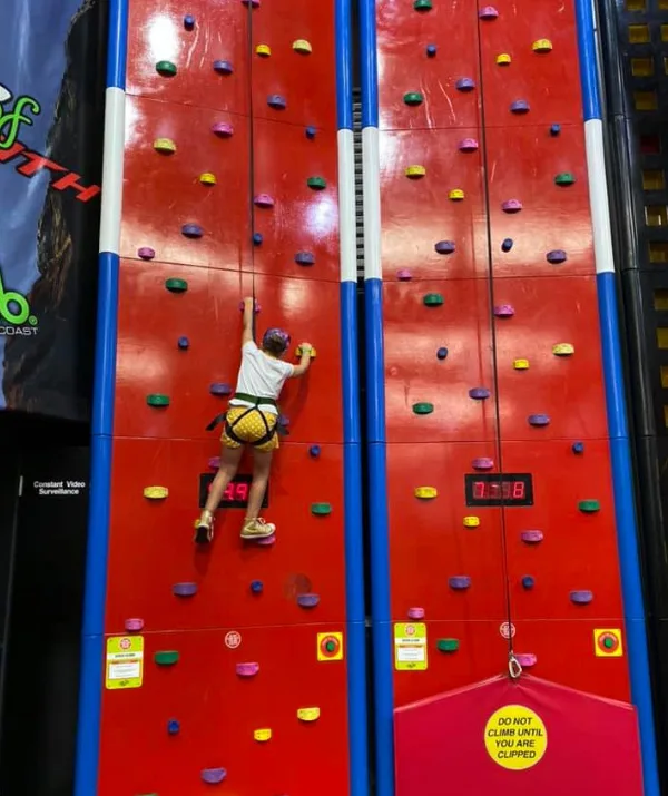 Person climbing a colorful indoor rock wall with a safety harness, timers visible.